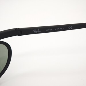 May include: Black Ray-Ban sunglasses with a matte finish. The temple arm features the Ray-Ban logo and "MADE IN ITALY" text. The lenses are a muted green colour.