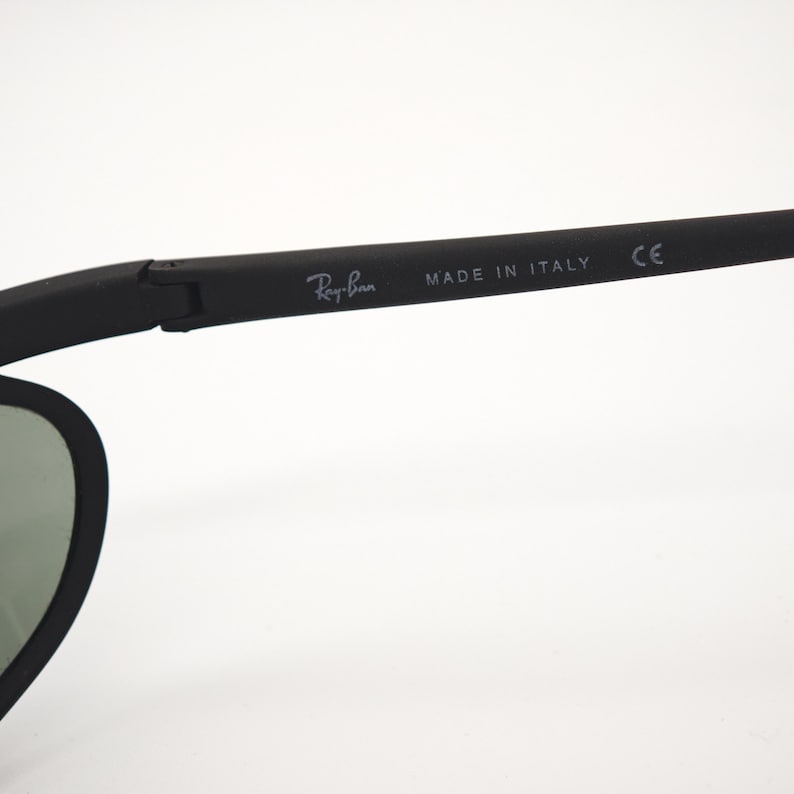 May include: Black Ray-Ban sunglasses with a matte finish. The temple arm features the Ray-Ban logo and "MADE IN ITALY" text. The lenses are a muted green colour.