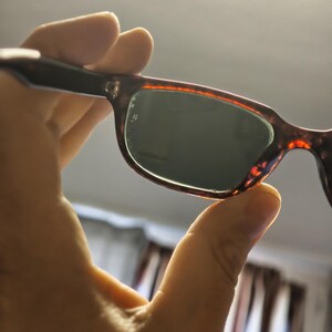 May include: Sunglasses with a tortoiseshell frame and dark lenses are held up. The frame has a brown and orange pattern. The lenses are a dark green color. The sunglasses are held up by a hand.