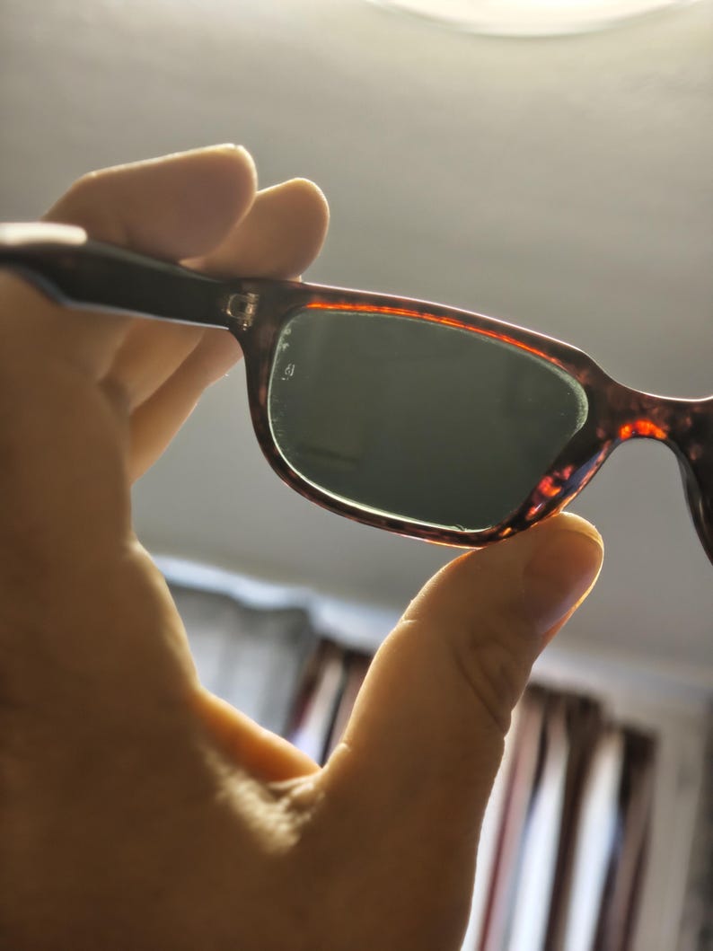 May include: Sunglasses with a tortoiseshell frame and dark lenses are held up. The frame has a brown and orange pattern. The lenses are a dark green color. The sunglasses are held up by a hand.