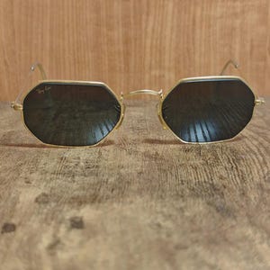 May include: Gold-framed sunglasses with dark lenses. The sunglasses have an octagonal shape and the Ray-Ban logo is visible on the upper left lens. The glasses are set against a wooden background.