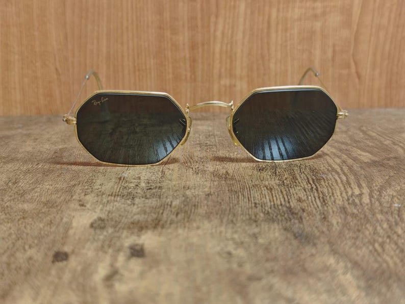 May include: Gold-framed sunglasses with dark lenses. The sunglasses have an octagonal shape and the Ray-Ban logo is visible on the upper left lens. The glasses are set against a wooden background.