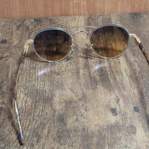 Peut inclure: Lunettes de soleil rondes avec une monture en m&eacute;tal dor&eacute; et des verres marron d&eacute;grad&eacute;s. Les branches pr&eacute;sentent un motif &eacute;caille de tortue. Les lunettes reposent sur une surface en bois.