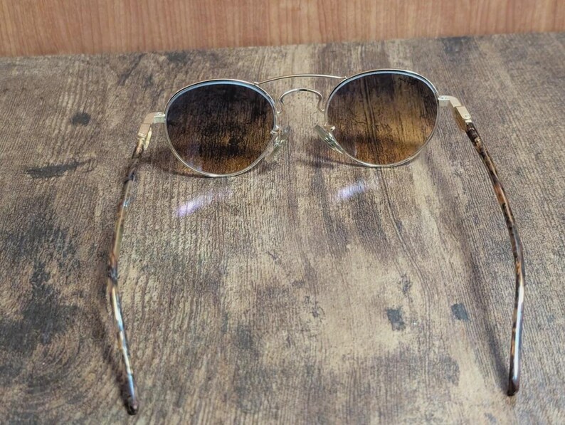 Peut inclure: Lunettes de soleil rondes avec une monture en m&eacute;tal dor&eacute; et des verres marron d&eacute;grad&eacute;s. Les branches pr&eacute;sentent un motif &eacute;caille de tortue. Les lunettes reposent sur une surface en bois.