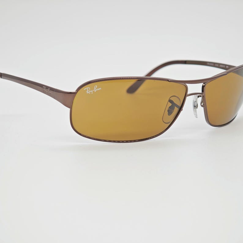 May include: Brown Ray-Ban sunglasses with a rectangular frame and amber-colored lenses. The Ray-Ban logo is visible on the left temple and lens. The sunglasses have a sleek, modern design.