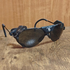 Può includere: Occhiali da sole neri con lenti scure e protezioni laterali in pelle. Gli occhiali hanno una montatura curva e il marchio "Julbo" è visibile sul lato. Progettati per attività all'aperto.