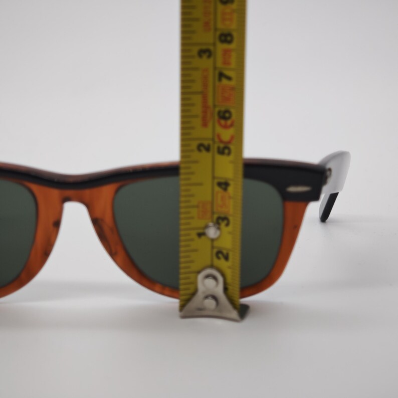 May include: Sunglasses with a brown wood-grain frame and dark lenses. The top of the frame is black. A yellow tape measure is placed over the sunglasses, showing a measurement of approximately 3 inches.