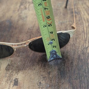 May include: Gold-framed sunglasses with dark lenses are measured with a tape measure. The tape measure shows a measurement of approximately 2 inches. The sunglasses are on a wooden surface.