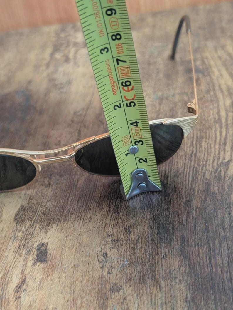 May include: Gold-framed sunglasses with dark lenses are measured with a tape measure. The tape measure shows a measurement of approximately 2 inches. The sunglasses are on a wooden surface.