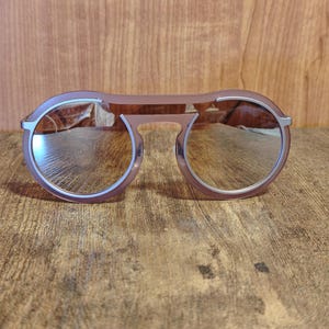 May include: Round-framed sunglasses with a mauve-colored frame and silver mirrored lenses. The sunglasses have a double bridge design and a keyhole nose bridge. The lenses are reflective, and the frame is a solid color.