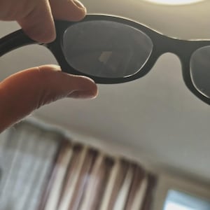 May include: Black sunglasses with dark lenses. The sunglasses have a classic, rectangular frame design. The image shows the sunglasses held up against a bright background, highlighting the shape and style of the eyewear.