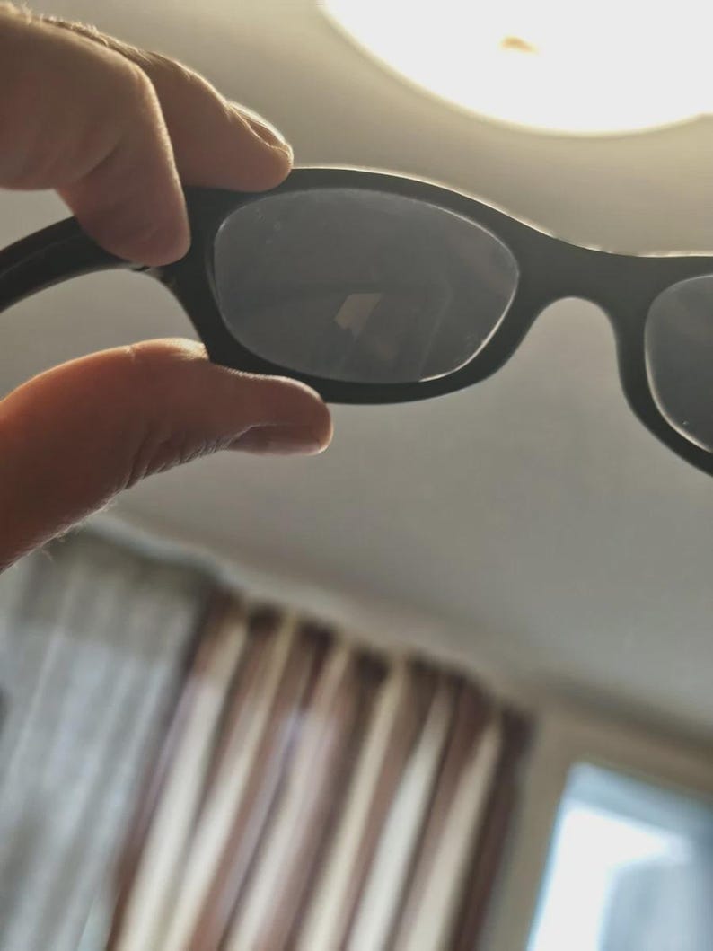 May include: Black sunglasses with dark lenses. The sunglasses have a classic, rectangular frame design. The image shows the sunglasses held up against a bright background, highlighting the shape and style of the eyewear.