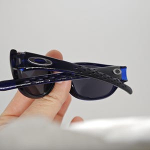 May include: Dark blue sunglasses with black lenses. The arms of the sunglasses have a textured design and the Oakley logo is visible on the temples. The sunglasses are held in a hand.