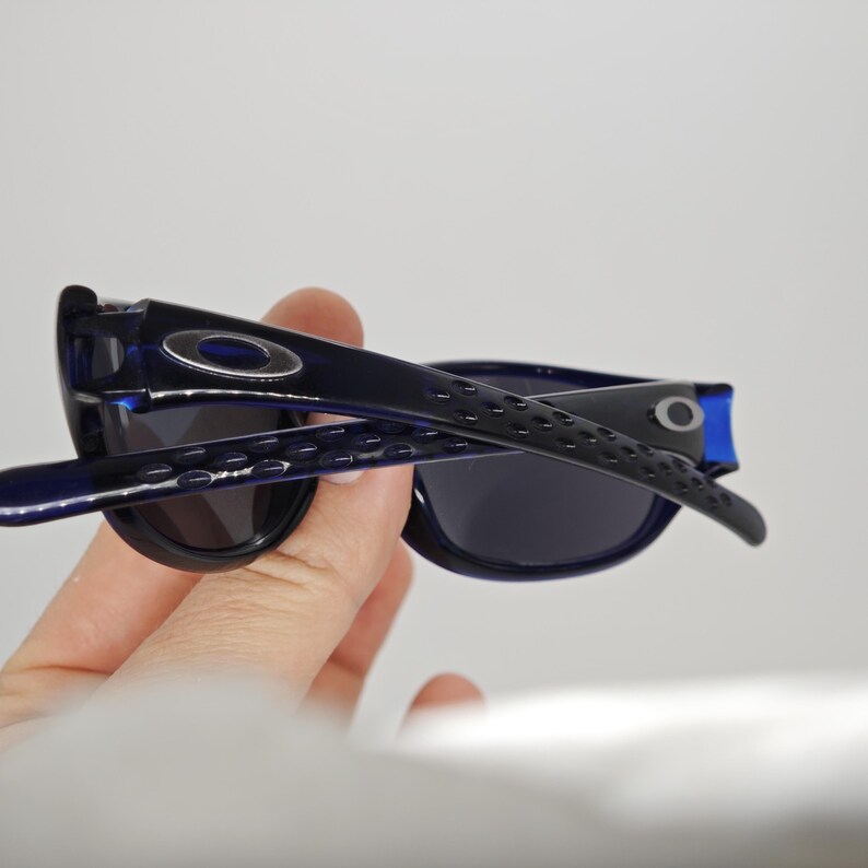 May include: Dark blue sunglasses with black lenses. The arms of the sunglasses have a textured design and the Oakley logo is visible on the temples. The sunglasses are held in a hand.