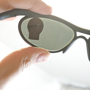 May include: Black sunglasses with green-tinted lenses. The sunglasses have a modern design with a matte finish. The frame is held by a hand, showing the shape and style of the eyewear. The lenses are oval-shaped.