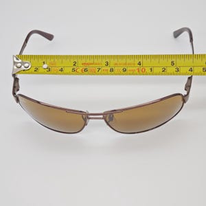 May include: Brown sunglasses with a brown frame and lenses. The sunglasses are measured with a yellow tape measure, showing a width of approximately 5 inches. The glasses have a sleek, modern design.