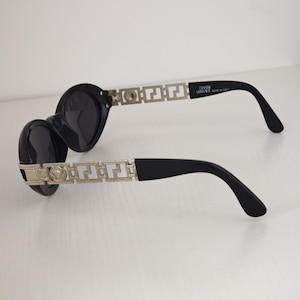 Puede incluir: Gafas de sol negras con lentes redondas y patillas de metal plateado con un patr&oacute;n geom&eacute;trico de greca y un emblema circular de Medusa. El interior de la patilla tiene impreso el texto GIANNI VERSACE MADE IN ITALY en letras blancas.