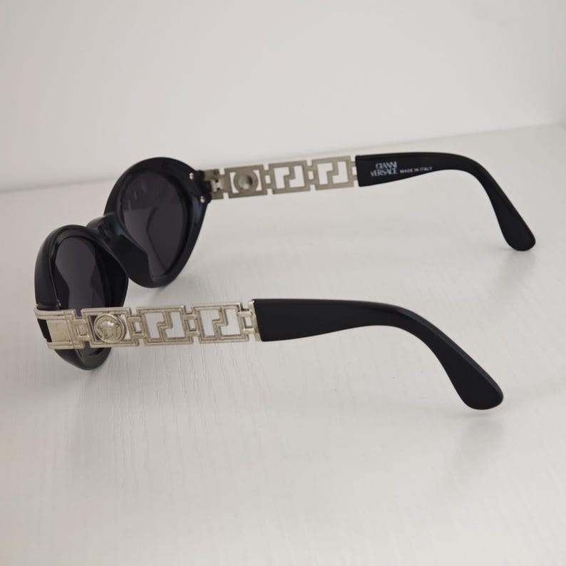 Puede incluir: Gafas de sol negras con lentes redondas y patillas de metal plateado con un patr&oacute;n geom&eacute;trico de greca y un emblema circular de Medusa. El interior de la patilla tiene impreso el texto GIANNI VERSACE MADE IN ITALY en letras blancas.