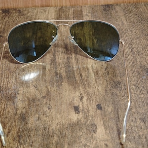 May include: Aviator sunglasses with a silver frame and dark lenses. The sunglasses are resting on a wooden surface. The lenses reflect the surrounding environment.