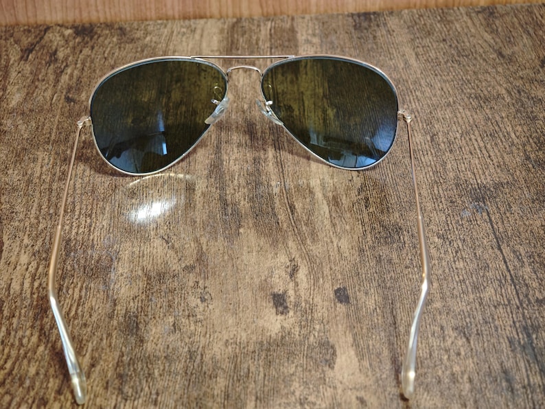 May include: Aviator sunglasses with a silver frame and dark lenses. The sunglasses are resting on a wooden surface. The lenses reflect the surrounding environment.
