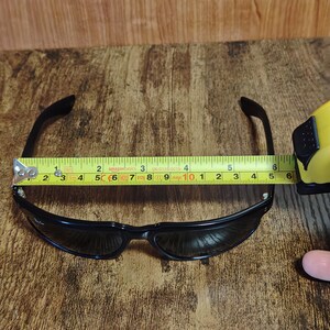May include: Black sunglasses with a curved frame, measured at approximately 6 inches wide. The sunglasses are positioned on a wooden surface, with a measuring tape placed across the frame.