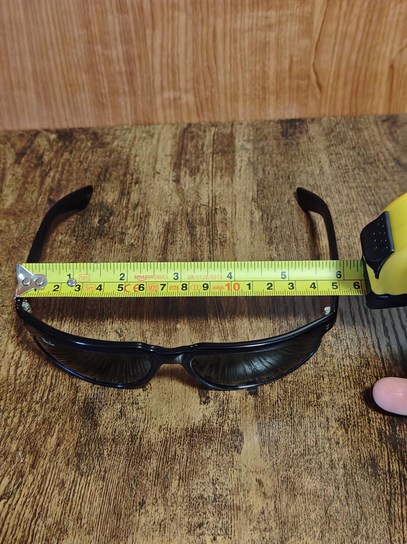 May include: Black sunglasses with a curved frame, measured at approximately 6 inches wide. The sunglasses are positioned on a wooden surface, with a measuring tape placed across the frame.