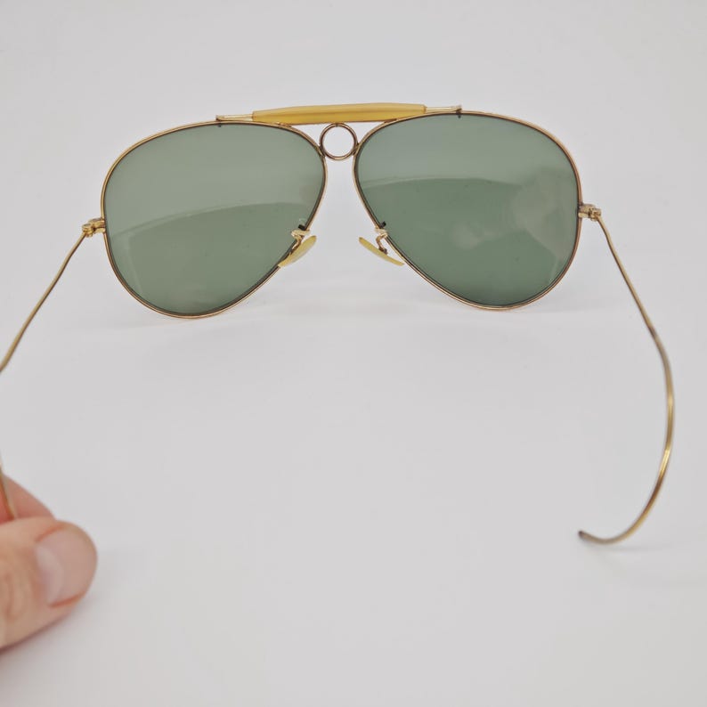 May include: Gold-framed aviator sunglasses with green lenses. The sunglasses have a double bridge and a light-colored bar across the top. The thin temples curve at the ends. The lenses are teardrop-shaped.