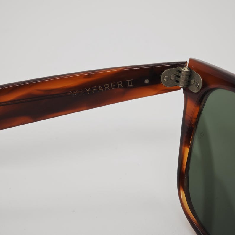 May include: Close-up of Wayfarer II sunglasses. The frames are a tortoiseshell pattern, with a dark green lens visible. The hinge is silver. The text "WAYFARER II" is printed on the arm of the glasses.