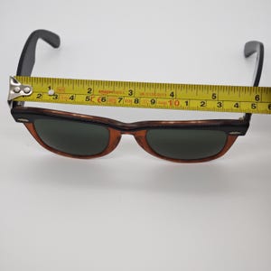 May include: Brown and black sunglasses with dark lenses. The sunglasses are measured with a yellow tape measure, showing a length of approximately 5.75 inches. The frames have a tortoiseshell pattern.