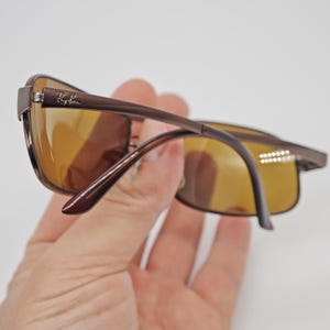 May include: Brown rectangular sunglasses with amber-colored lenses. The sunglasses have a thin, dark brown frame and the Ray-Ban logo is visible on the temple. The lenses are a warm, amber color.