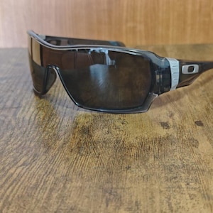 May include: Black sunglasses with a wraparound design. The sunglasses have a dark lens and a black frame with a silver accent on the temples. The sunglasses are resting on a wooden surface.