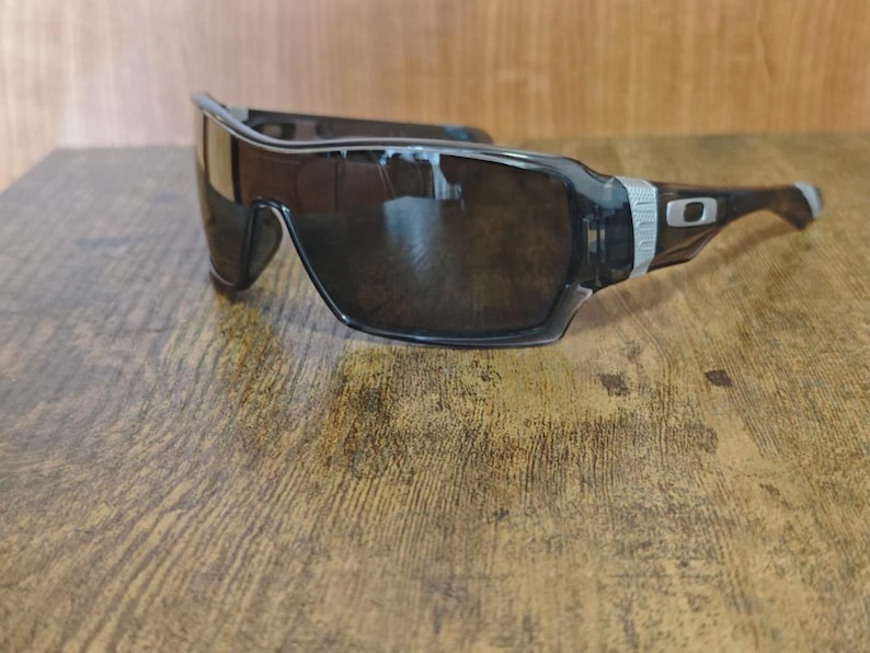 May include: Black sunglasses with a wraparound design. The sunglasses have a dark lens and a black frame with a silver accent on the temples. The sunglasses are resting on a wooden surface.