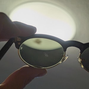 May include: Black and gold-rimmed sunglasses with green lenses. The sunglasses have a classic, rounded shape and are held up against a bright light source. The frame is black with gold accents.