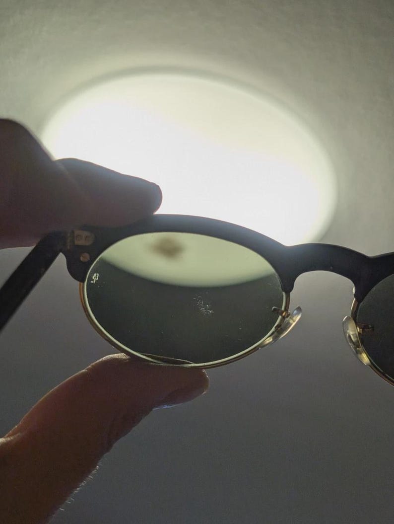 May include: Black and gold-rimmed sunglasses with green lenses. The sunglasses have a classic, rounded shape and are held up against a bright light source. The frame is black with gold accents.