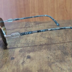 May include: A pair of sunglasses with a silver frame and dark lenses. The temples feature a blue and black marbled pattern. The glasses are resting on a wooden surface.