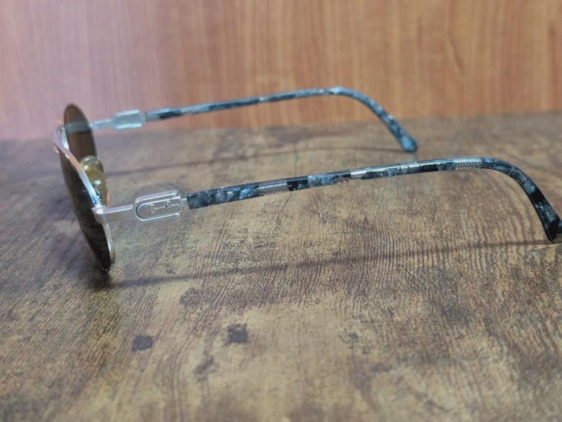 May include: A pair of sunglasses with a silver frame and dark lenses. The temples feature a blue and black marbled pattern. The glasses are resting on a wooden surface.