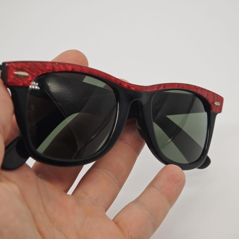May include: Black sunglasses with a red patterned frame. The sunglasses have dark lenses and are held in a hand against a white background. The sunglasses are a classic style.