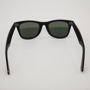 May include: Black sunglasses with dark green lenses. The sunglasses have a classic rectangular frame design. The arms of the sunglasses are extended, showing the hinges and the shape of the frame.