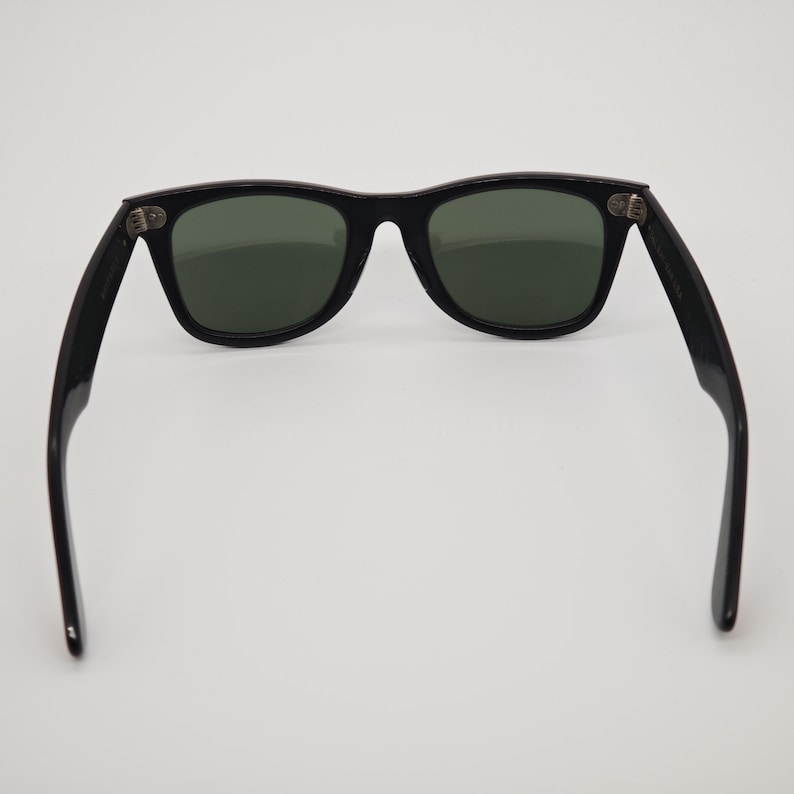 May include: Black sunglasses with dark green lenses. The sunglasses have a classic rectangular frame design. The arms of the sunglasses are extended, showing the hinges and the shape of the frame.