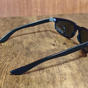 May include: Black sunglasses with dark lenses. The sunglasses have a classic rectangular frame and sleek, straight arms. The lenses are reflective, and the sunglasses are resting on a wooden surface.