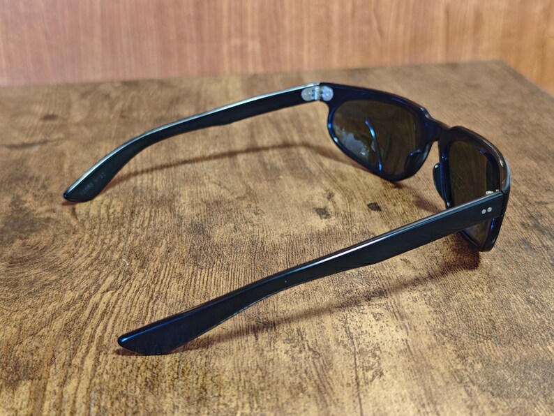 May include: Black sunglasses with dark lenses. The sunglasses have a classic rectangular frame and sleek, straight arms. The lenses are reflective, and the sunglasses are resting on a wooden surface.