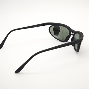 May include: Black sunglasses with a curved frame and oval lenses. The sunglasses have a matte finish and a sleek design. The lenses are a dark green colour. The text "FS5 W2172" is printed on the arm.