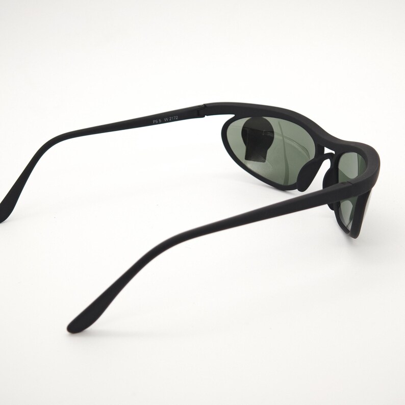 May include: Black sunglasses with a curved frame and oval lenses. The sunglasses have a matte finish and a sleek design. The lenses are a dark green colour. The text "FS5 W2172" is printed on the arm.