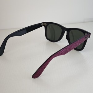 Puede incluir: Gafas de sol negras con lentes de color verde oscuro y un patr&oacute;n morado texturizado en las patillas exteriores. La montura tiene una forma cl&aacute;sica con bisagras de metal plateado y el logotipo de Ray-Ban es visible en el lateral.