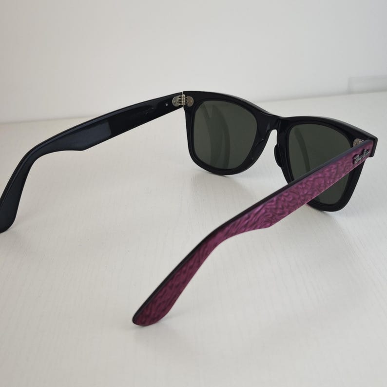 Puede incluir: Gafas de sol negras con lentes de color verde oscuro y un patr&oacute;n morado texturizado en las patillas exteriores. La montura tiene una forma cl&aacute;sica con bisagras de metal plateado y el logotipo de Ray-Ban es visible en el lateral.