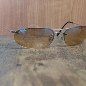 May include: Rimless sunglasses with a silver frame and brown gradient lenses. The Ray-Ban logo is visible on the upper left lens. The sunglasses have a classic, oval shape and are set against a wooden background.