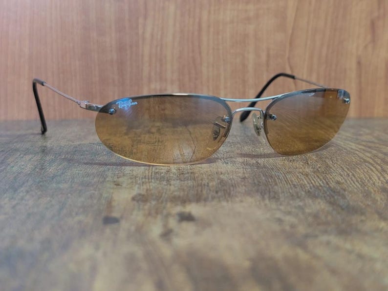 May include: Rimless sunglasses with a silver frame and brown gradient lenses. The Ray-Ban logo is visible on the upper left lens. The sunglasses have a classic, oval shape and are set against a wooden background.
