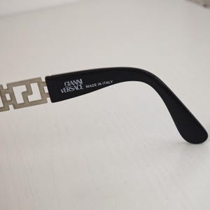Puede incluir: Una patilla negra de gafas con una bisagra de metal plateado con un patr&oacute;n de llave griega. El lado interior de la patilla negra tiene impreso el texto GIANNI VERSACE y MADE IN ITALY en letras blancas.