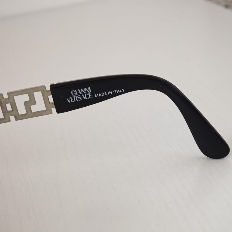 Puede incluir: Una patilla negra de gafas con una bisagra de metal plateado con un patr&oacute;n de llave griega. El lado interior de la patilla negra tiene impreso el texto GIANNI VERSACE y MADE IN ITALY en letras blancas.