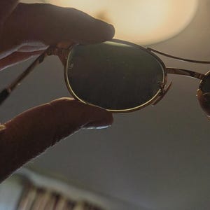 May include: Gold-rimmed sunglasses with dark green lenses. The sunglasses have a classic, round shape and a thin metal frame. The image is taken indoors, with a blurred background and a hand holding the sunglasses.
